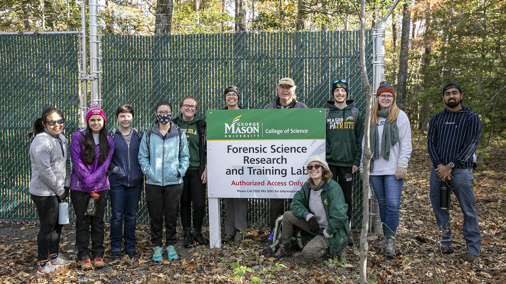 Ten students and professors stand beside a sign Forensic Sciences Research and Training Lab. It's fall, students are wearing jackets.