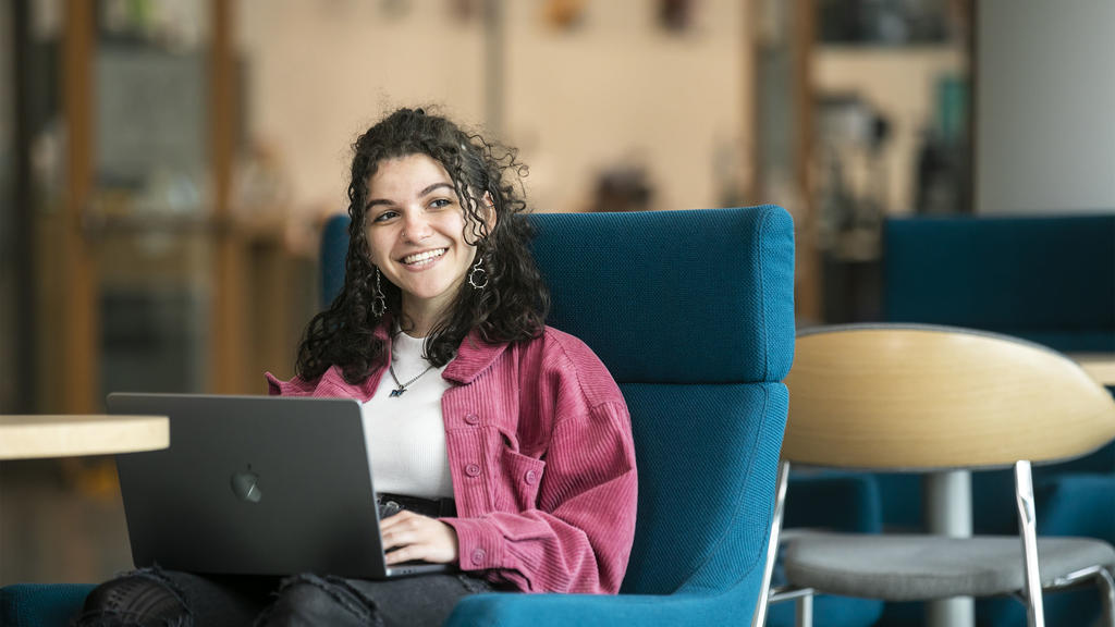 female student with long brown curly hair sits in a bright blue chair. She is looking at the camera, a laptop on her lap.