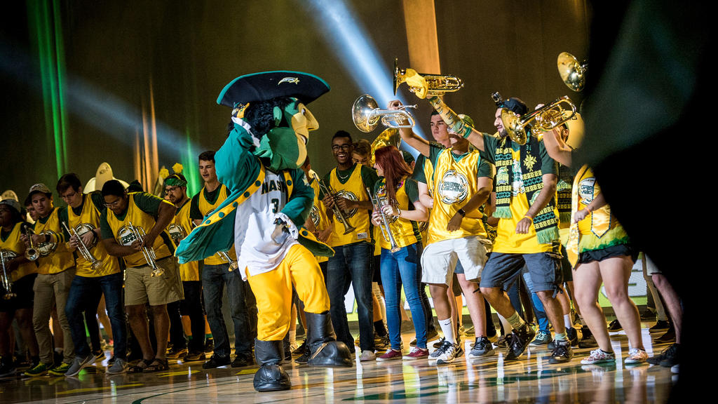 The Green Machine pep band plays in the EagleBank Arena. Members of the band dance with the Patriot mascot.