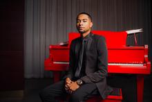 Deion Maith, dressed in a suit, sits on a red piano stool next to a red piano