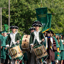 Three drummers in colonial garb of green and white march ahead of a procession of graduates in regalia.