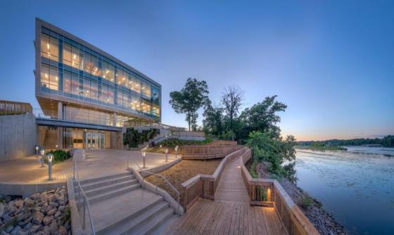 Photo of the Potomac Science Center, a three story building with large glass windows, sitting on the waterfront. There is a large concrete patio and a wooden deck. It is twilight and the building is illuminated against the sky