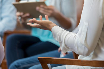 A woman gestures with her left arm, palm facing upward. Her face is not visible, she appears to be talking to a group in a talk therapy setting