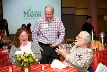 A group of three faculty and staff sit and stand together at a breakfast event on the Fairfax Campus. They are talking and laughing. The table is set with colorful linens, votives, and a flower arrangement. The presentation screen in the background has a George Mason University logo displayed.